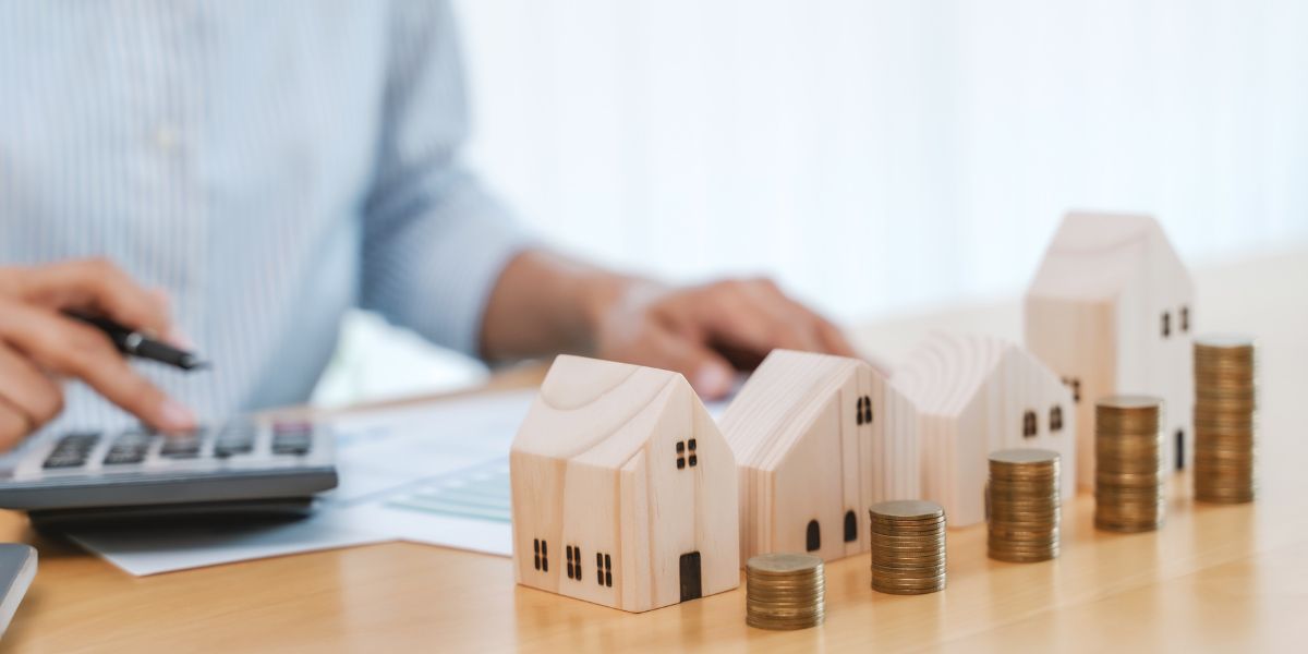 Person calculating a lien and back taxes next to small wooden house models and stacked coins.