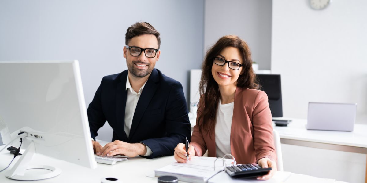 Two professionals smiling at a desk, reviewing documents and using a computer.