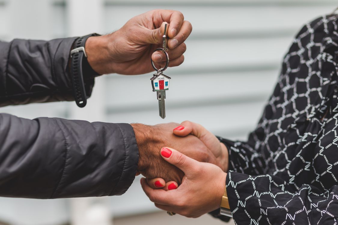 A close-up shot of a hand carefully placing a coin into a white, house-shaped piggy bank, symbolizing the savings and profit maximization from a home sale.
