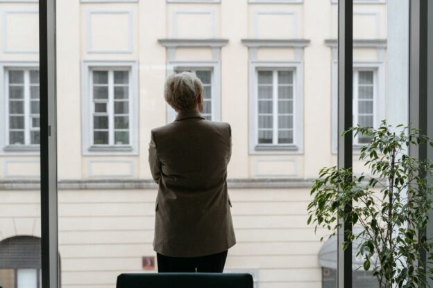 Professionally dressed man stands pensively, looking out the large window of a modern high-rise office, symbolizing the hi...