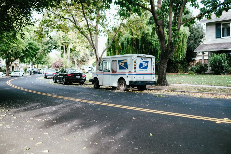 A delivery van drives down a quiet suburban street lined with houses, representing the 'last mile' of the e-commerce supply chain changing neighborhoods.